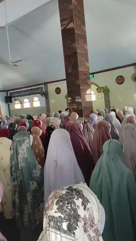 The video shows a large group of women in a mosque, dressed in colorful hijabs and prayer garments. They are standing in rows, facing the same direction, seemingly in the middle of a prayer service. The mosque appears to have high ceilings with exposed wooden beams and ceiling fans. A tall, marble-clad pillar is prominently featured in the center of the frame. The walls are adorned with Islamic calligraphy. Sunlight streams in from arched windows at the back of the mosque. The overall atmosphere suggests a communal religious gathering.