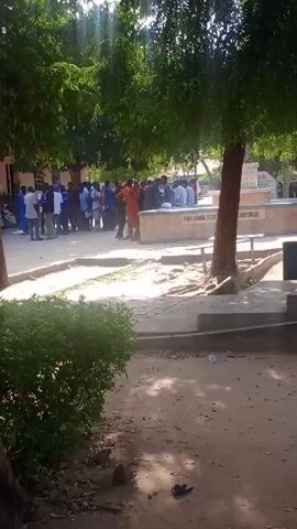 The video shows a partially obscured view of an outdoor gathering of people, likely students, near a building labeled "Prof. Lawal Audi Auditorium". There are several trees providing shade over the area, with sunlight filtering through the leaves. A person is briefly seen sitting on a ledge in the foreground, looking at a phone. The overall impression is of a casual, possibly academic, environment.