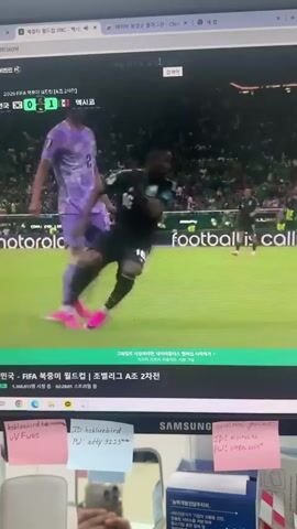 A computer monitor displays a live soccer match. The score is displayed as South Korea 0, Mexico 1. The match appears to be part of the FIFA World Cup. The lower portion of the screen shows a text overlay in Korean. There are sticky notes attached to the monitor bezel, with text in English. One note reads "ID: hshluebird PJ: adty 9223" and another reads "ID: milnakal PJ: open-copy". The monitor is placed on a desk, with a blue box and some papers visible in the lower right corner.