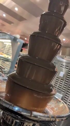 The video shows a chocolate fountain. It is a four-tiered fountain with chocolate flowing down each tier. The fountain is made of stainless steel and has a shallow circular base. There are various foods available to dip in the chocolate, including fruits and marshmallows, on a nearby buffet. The environment appears to be inside a restaurant or dining hall.