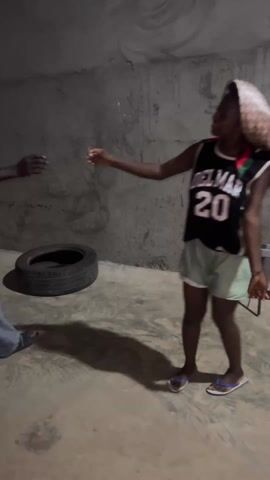 A woman wearing a headscarf, basketball jersey, and shorts is dancing in a concrete room. She dances solo, at times interacting with a man who offers his hand. She spins, moves her arms and legs, and smiles. The room is bare except for a tire, a couple of metal benches, and some papers taped to the wall.
