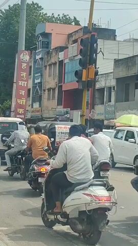 The video shows a street scene with moderate traffic. Several people are on motorcycles and scooters. One person is on a bicycle. There are cars and a small auto-rickshaw. Buildings line the street. There is a traffic signal and a tall pole with wires. A sign with writing in a non-English script is visible on the side of a building. The weather appears to be clear.