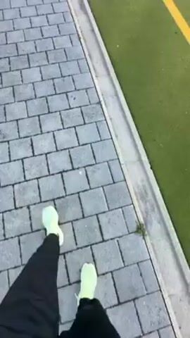 The video shows a person walking towards a green turf sports field on a sunny day. The person is wearing black pants and neon green sneakers and is walking along a brick pathway. To the right of the pathway is a strip of green turf with a yellow line marking the edge of the field. The camera pans to show the whole field, where several people dressed in green are standing and walking. There are also white cones and small blue markers on the field. A building and trees are visible in the background under a clear blue sky.