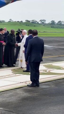 In an outdoor scene at what appears to be an airport tarmac in Malabo, Equatorial Guinea, Pope Francis, dressed in his white cassock, is seen disembarking from an airplane, with part of its wing visible in the upper left. He is being formally greeted by a line of officials and clergy.

The Pope is shaking hands with several men. One man in a black cassock with a purple stole shakes the Pope's hand, while another man in a similar black cassock waits nearby. Further down the line, a man in a dark suit also shakes the Pope's hand. Another man in a dark suit stands with his back to the camera, observing the greetings. A cameraman is visible in the background, filming the event.

The group stands on a patterned, light-colored walkway laid out on the tarmac, which appears wet, suggesting recent rain or an overcast day. In the background, there are lush green fields and trees under a cloudy sky. The atmosphere is formal and respectful.