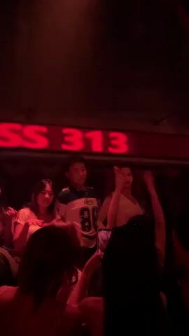 Three people are on a stage or raised platform in front of a neon sign that reads "CLASS 313". The person in the center, a male wearing a white and blue jersey with the number 86, is dancing and making gestures. To his left is a female with dark hair, wearing a light-colored top, who appears to be smiling and looking at the male. To the male's right is another female, also with dark hair, wearing a white top, who is dancing with her hands raised. In the foreground, partially obscuring the stage, are the backs of several people in the audience. One person in the audience is holding up a phone to record the performance. The environment is dimly lit with red and purple lighting.