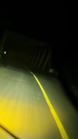 This is a first-person perspective video, likely from a car, driving through a tunnel at night. The headlights illuminate the asphalt road with a yellow glow, highlighting a bright yellow dividing line. The tunnel structure is visible on the sides and overhead, characterized by dark, angled supports and a low ceiling, creating a sense of enclosure. The darkness outside the illuminated path suggests the tunnel is long and deep. There are no other vehicles or people visible in the immediate vicinity.