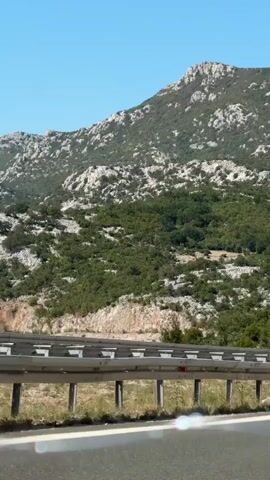 A first-person perspective video shows a view from a moving vehicle traveling on a highway. In the foreground, the asphalt road with white lane markings is visible. A metal guardrail separates the road from the adjacent terrain. Beyond the guardrail, dry grass and rocky ground are seen. The background is dominated by a large, sparsely vegetated mountain range with rocky outcrops. The sky is clear and blue. As the vehicle moves, other vehicles are visible on the highway, including a white van with the word "DOTA" on its side, a truck, and a blue passenger car. The terrain along the highway includes rock cuts and some scattered trees and bushes. The lighting suggests it is daytime with bright sunlight.