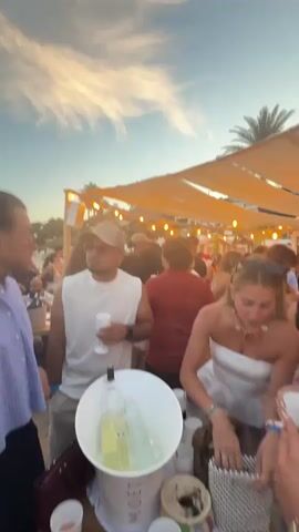 The video depicts an outdoor party scene at sunset, with a clear sky displaying wispy clouds and warm hues. A diverse group of adults, dressed in casual and semi-formal attire, are mingling and socializing. In the foreground, a man in a white tank top and baseball cap holds a white cup, standing near a large white bucket filled with bottles chilling in ice. People are actively talking, laughing, and holding drinks. Canopies adorned with string lights are visible overhead, and palm trees are present in the background. The camera pans across the crowd, capturing various groups interacting in a lively atmosphere, with the ocean visible in the distance.