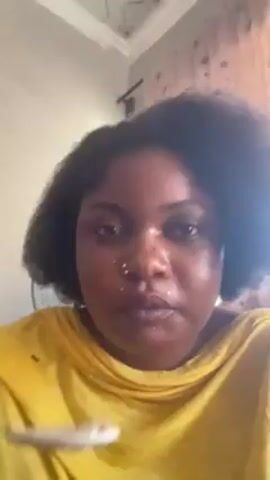 A woman with dark hair and a yellow shirt is sitting indoors and eating something with a spoon. She looks up and around after taking a bite. There are small white specks on her face and shirt, likely from the food. The background includes a wall with some decorative elements.