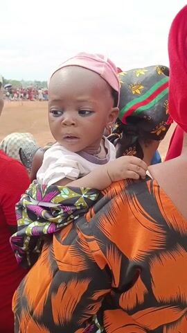 A close-up shot shows a baby being carried on the back of an adult woman. The baby, wearing a light pink cap, a white top, and a colorful wrap, looks to the left with a slightly worried expression. The woman carrying the baby wears an orange and black patterned top and a red headscarf. Another woman beside them is wearing a similar headscarf. A crowd of people is visible in the background on a dirt field under a cloudy sky.