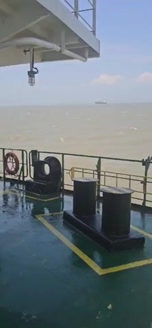 A video captures a view from the deck of a ferry, looking out towards the ocean. The deck is wet, likely from rain or sea spray, and marked with yellow safety lines. Several pieces of marine equipment are visible, including a large, black bollard with a curved opening and two black cylindrical bollards on a raised platform. A life preserver hangs on the railing to the left. In the distance, a cargo ship is visible on the horizon under a hazy sky with scattered clouds. The water appears choppy, suggesting a moderate sea state.