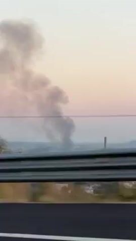 The video shows a view from a moving vehicle, likely a car, driving alongside a highway. A gray guardrail is visible in the foreground. Beyond the guardrail, a field of brown vegetation is present. In the distance, a column of gray smoke rises against a pale sky with a hint of pink. The movement of the car causes blurring of the landscape.