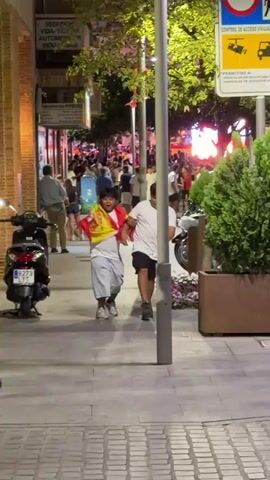 A young person wearing a white t-shirt and light blue shorts, draped in a Spanish flag, runs down a sidewalk. They are looking back over their shoulder and appear to be laughing. Two other young people, one wearing a red shirt and the other a white t-shirt, are running behind them. In the background, a crowd of people are visible, as well as buildings and trees. The scene is outdoors at night.