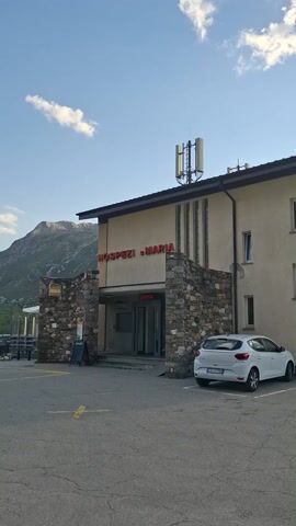 This media file depicts a scenic mountain landscape in Medel (Lucmagn), Switzerland, likely during the daytime given the bright sunlight. The scene features a building complex, possibly a hotel or guesthouse, with a facade of light-colored bricks and stone accents. A white car is parked in front of the building, and another car is partially visible in the background.

The surrounding environment is characterized by rugged, verdant mountains under a partly cloudy sky. The sunlight casts shadows and highlights the contours of the terrain, suggesting a clear or fair weather day. A road runs alongside the mountainside, indicating a likely tourist or transit route.

Visible text on the building includes "OSPEZI MARIA" in large red letters and "Uscita" above a doorway, which is Italian for "Exit." The white car also has text on its side, which appears to be "TERRADATA" followed by smaller text below that reads "MESSEN SE DOKUMENTATION."

The overall impression is one of a serene, mountainous location, possibly a stopover for travelers or visitors seeking nature and relaxation. The combination of the man-made structures and the natural grandeur creates a sense of both human presence and awe-inspiring wilderness. The media file captures a moment of stillness, with no explicit activity or people clearly visible, allowing the viewer to appreciate the quiet beauty of the location.