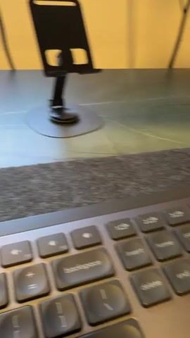 The camera focuses on a gray keyboard with black keys. In the background, there is a black smartphone holder on a dark surface. The background wall is a pale yellow.