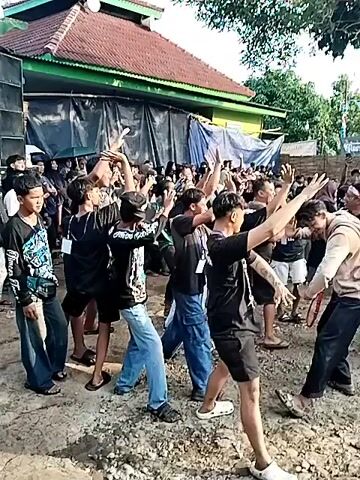 A crowd of young men are gathered in front of a building that has a green roof. They are dancing and clapping along to music being played from a large speaker on the left.  Most of the men are wearing black shirts.  Some of them are wearing headbands. Someone throws water on the crowd. There are several trees around the crowd.