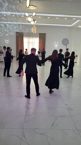 The video shows a group of people in formal black attire dancing in a large, well-lit room with a polished floor. A couple in the foreground is dancing closely together, facing away from the camera. Other individuals are scattered around the room, also dancing or standing. In the background, there are tables and chairs, suggesting a formal event like a wedding reception or a gala. The room is decorated with modern light fixtures. The overall atmosphere appears festive and celebratory.