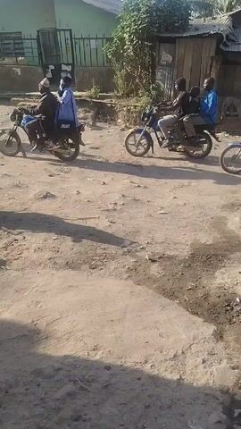 The video shows a dirt road with several motorcycles riding by. Some of the motorcycles carry multiple passengers. There is a parked motorcycle on the side of the road. Buildings are visible in the background. The area appears to be a residential neighborhood. A trench with debris is visible on the side of the road. The video is taken during the daytime.
