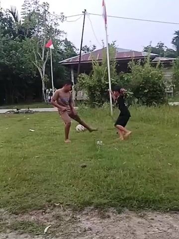 Two young boys are playing soccer in a grassy field. One boy is wearing a brown t-shirt and shorts, while the other is wearing a black t-shirt with a white number on the back and shorts. They are both barefoot. There is a soccer ball on the ground between them. In the background, there are trees, a flag, and a building. The flag appears to be the Indonesian flag. The overall scene suggests a casual outdoor activity.