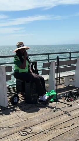 A woman wearing a straw hat with a green and white band, a green and black striped shirt, and black pants sits on a black portable speaker. She is holding a microphone to her mouth and appears to be singing or speaking into it. In front of her, on the wooden pier, is a microphone stand with a microphone attached, and a pile of green fabric. Behind her, there are white benches and a railing overlooking the ocean. The sky is blue with some white clouds. The scene is lit by daylight.
