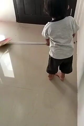A young child with short black hair, wearing a gray t-shirt and dark shorts, stands barefoot on a light-colored tile floor and sweeps with a small broom that has a red and beige head and a wooden handle. The child sweeps the floor towards a dark brown door. The room has white walls and natural light is visible in the reflection on the tiled floor.