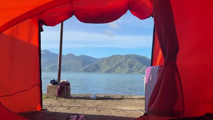 The video shows a view from inside an orange tent. The tent flap is open, revealing a body of water, possibly a lake, and mountains in the background. The sky is blue with some clouds. On the ground outside the tent, there are some objects, including a purple bag, a water bottle, and a pair of flip-flops. The overall scene suggests a camping or outdoor recreational setting. The camera appears to be stationary, capturing a static view of the environment. There are no people or animals visible in the frame. The lighting indicates it is daytime, possibly morning or afternoon.