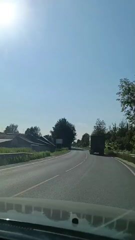 The video captures a view from inside a moving vehicle, looking forward. The scene is a rural road on a bright, sunny day with a clear blue sky. The sun is prominently visible in the upper portion of the frame, casting a bright glare. On the left side of the road, there are some low buildings with tiled roofs and vegetation. Further down the road, there are trees and bushes lining the side. A large truck is visible in the distance, also moving along the road. The road itself appears to be paved with white lines marking the lanes. The overall environment suggests a journey through a countryside area.