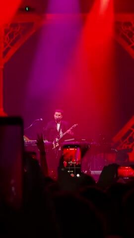 A man in a suit is playing a guitar on a stage under pink and red lights. He is standing in front of a keyboard and other musical equipment. In the foreground, there are many hands holding up phones to record the performance. The stage has decorative red arches on the sides.