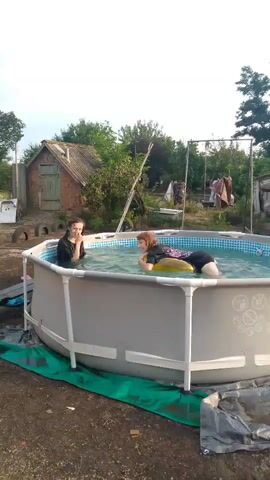 A still shot from a video shows a backyard scene with an above-ground swimming pool. Two young women are in the pool, both wearing dark-colored swimwear. One woman is on the left side of the pool, facing forward and looking towards the camera. She has long dark hair and her hands are submerged in the water. The other woman is on the right side of the pool, lying on a yellow inflatable object, possibly a ring or pillow. She has reddish-brown hair and is also looking towards the camera. The pool is filled with clear water and has a checkered pattern around the inside rim. The pool is set on a dirt surface and is surrounded by a green tarp on the ground. In the background, there is a small, rustic brick building with a wooden door and a sloped roof. Trees and bushes are visible behind the building, and a swing set with two swings is also present. The sky is overcast, suggesting a cloudy day. The overall impression is a casual, outdoor setting.