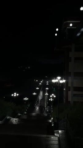 The video shows a nighttime scene of a wet, illuminated pathway lined with streetlights. The pathway appears to be a gently sloping walkway or bridge, with a set of stairs leading down to it from the foreground. Multiple streetlights with multiple globes are visible along both sides of the pathway, casting a bright glow on the wet surface and creating reflections. In the distance, more lights can be seen, suggesting the pathway extends further. A few figures, seemingly people, are walking along the illuminated pathway in the mid-ground, their forms silhouetted against the lights. To the right of the pathway, a building with a distinctive architectural style, featuring horizontal banding and illuminated upper sections, is partially visible. The environment appears to be outdoors, possibly a park or waterfront area, given the presence of trees or shrubbery bordering the pathway. The overall atmosphere is dark, with the artificial lights providing the primary illumination.
