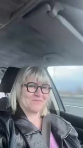 The video shows a middle-aged woman with short blonde hair and glasses, wearing a black jacket and a pink top, sitting in the driver's seat of a car. She is looking at the camera and making faces. The car is moving on a highway, with other vehicles visible outside the window. A water tower painted with blue and yellow camouflage is seen in the distance. The interior of the car is mostly gray. There is a headrest behind the driver and a sun visor above. The sky is overcast. Audio of someone speaking is also present.