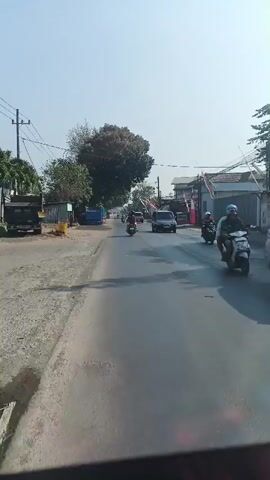 A sunny daytime shot from a moving vehicle, looking down a paved road lined with buildings and trees. Several motorcycles and a few cars are driving on the road. Some of the buildings have Indonesian flags displayed. There is a blue truck parked on the side of the road. The sky is clear and blue.
