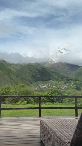 The video shows a static, wide-angle shot of a mountainous landscape. In the foreground, a wooden deck with a wicker lounge chair is visible. A wooden railing separates the deck from a grassy area, beyond which lush green hills rise. In the mid-ground, a village with red-roofed buildings is nestled in a valley. The background is dominated by a majestic snow-capped mountain peak partially obscured by clouds. The sky is blue with scattered white clouds. The lighting suggests daylight, possibly late morning or early afternoon. The camera appears to be stationary, and no people or discernible actions are taking place. The overall atmosphere is serene and natural.