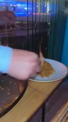 A person plates a small serving of a paella-like dish with a single large shrimp on top. The dish is placed on a white plate with a speckled gold rim, alongside other plates of the same dish. A large, used paella pan sits to the left. The plating is done on a wooden table next to a window.