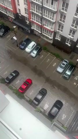 The video shows a top-down view of a parking lot next to an apartment building. Several cars are parked in designated spots, including a red car, a black car, a white car, a silver car, and a blue car. The parking lot appears to be wet, suggesting it may have rained recently. The apartment building has multiple floors with numerous windows. There are also some plants and trees along the edge of the parking lot. No people are visible in the video.