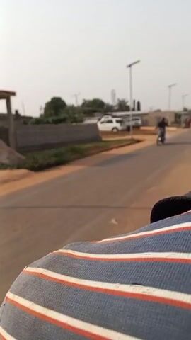 The video shows a street in a town. It is daytime and the sky is bright. The camera is moving, likely from a vehicle. The street is paved, and there are sidewalks on either side. Several buildings are visible, including houses, shops, and a car wash area with a few vehicles parked there. There are also some piles of construction material, such as bricks and lumber, along the roadside. In one scene, two people are walking along the street. There are also several streetlights lining the road. One scene shows a motorcycle passing.