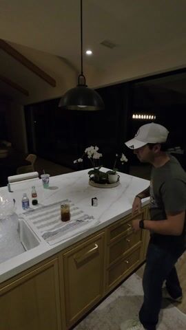 The video shows a group of young men gathered around a kitchen island. The island has a white countertop and light wood cabinets. On the countertop, there is a bag of ice in the sink, drinks, alcohol bottles, limes, a knife, and other items. Four young men are visible in the video: one in a white baseball cap and a green shirt, one in a gray hoodie, one in a blue sweatshirt, and one in a tank top. The young men are standing around the kitchen island; one is looking at his phone. The room is well-lit by recessed lighting and a pendant light hanging over the island. In the background, there is a dining area and living room visible through a large window. A staircase is visible in the background.