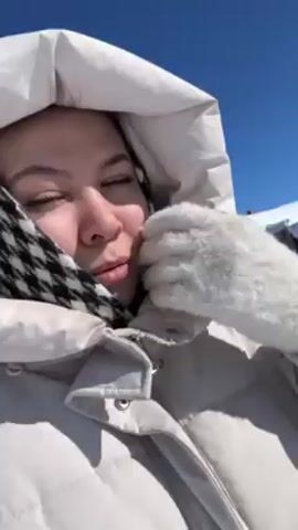 A person wearing a light-colored puffy jacket with a hood and a black and white patterned scarf is filming a video of themselves in a snowy environment. They are smiling and appear to be enjoying the outdoors. The sky is clear and blue. In the background, there are snow-covered buildings and trees. The person adjusts the hood of their jacket and continues to film themselves while walking through the snow.