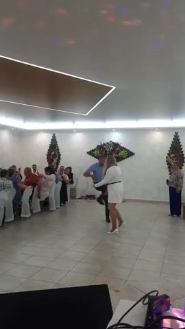 This video shows a wedding reception. A man and a woman are dancing in the center of the room. There are tables with chairs set up on the left side of the room, and people are seated at the tables watching the dancers. There are also some decorations on the walls, and disco lights are visible on the ceiling. The floor is tiled, and the room appears to be well-lit.
