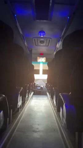 The media file depicts the interior of a bus, looking forward towards the windshield. The aisle of the bus is visible, with rows of dark-colored seats on either side. The seats appear to be empty. The ceiling of the bus is lit with blue LED lights. A ventilation unit is visible on the ceiling, along with a small screen displaying what looks like a television program. The windshield shows a road stretching into the distance, with a bright light emanating from beyond it, suggesting either sunrise, sunset, or a strong light source. The overall mood is quiet and still, with no visible activity or passengers. The scene is set during daytime, as indicated by the bright light outside. The location context provided is Шешенқара, Kazakhstan, but there are no specific visual cues in the media file to confirm this location. The visible text on the screen appears to be "2023", likely indicating the year.