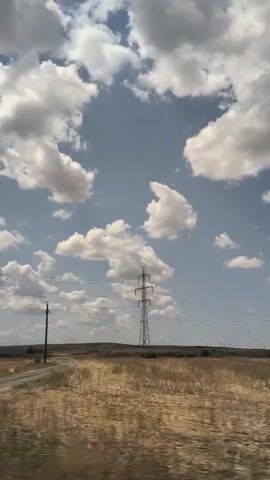 The video pans across a vast, open landscape under a bright blue sky with scattered white clouds. The ground is covered in dry, yellowed grass, suggesting a rural or agricultural area. A dirt road runs through the scene, leading towards a distant horizon. On the right side of the frame, a tall, metal electricity pylon stands prominently, with wires stretching across the sky. To the left of the pylon, a much shorter, dark pole with wires is visible. The overall impression is one of a dry, sparsely populated environment with infrastructure for power transmission.