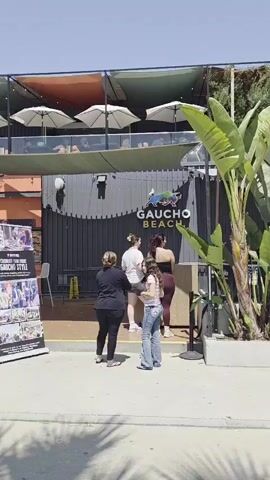 The video shows the exterior of a building with a sign that reads "GAUCHO BEACH". Four people are standing in front of the building. The person on the far left is wearing a dark shirt and dark pants. The person next to them is wearing a white shirt and dark pants. The third person is wearing a white top and blue jeans. The person on the far right is wearing a black top and maroon pants. There is a large green plant on the right side of the frame. Above the building, there are several umbrellas and people on a balcony. The weather appears to be sunny.