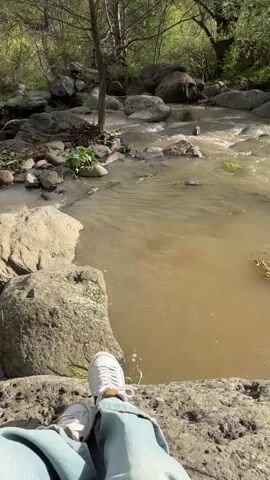 The video shows a first-person perspective of someone sitting on a rock next to a river. The river is flowing and the water is murky. There are several rocks and trees along the riverbank. A man walks across the rocks in the background. The person in the foreground is wearing white sneakers and light blue jeans.