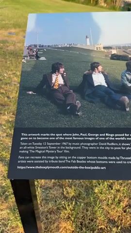 A static shot shows a sign that commemorates the spot where the Beatles took a famous photo. The sign features the original photo, showing John, Paul, George, and Ringo reclining on the grass, with Smeaton's Tower visible in the background. Text on the sign explains that the photo was taken on September 12, 1967, by photographer David Redfern. It also mentions that fans can recreate the image by sitting on copper bottom molds made by Thrussell and Thrussell. The sign includes a QR code and the Plymouth City Council logo. The sign is placed on a black metal frame, and the background consists of a grassy area. The sun is shining.