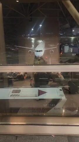 The video shows an airport gate area at night. A white airplane is parked on the tarmac outside the window. In the foreground, a portion of a baggage cart with the markings "UA0143" and "UTG" is visible. Passengers are seated in the gate area, and some appear to be standing near the window, looking out at the plane. The environment appears to be a modern airport terminal with a glass window separating the interior from the exterior tarmac. The lighting suggests it is nighttime, with artificial lights illuminating the tarmac.