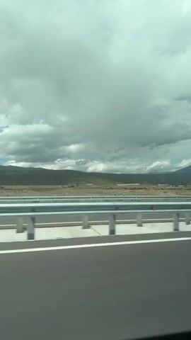 The video depicts a roadside view from a moving vehicle, likely a car or bus, traveling on a highway or bridge. The foreground consists of the asphalt road surface with lane markings, and a metal guardrail. Beyond the guardrail, there is a grassy plain with scattered buildings or structures in the middle distance. The background features rolling hills or mountains under a cloudy sky. The overall scene suggests travel through a rural or semi-rural area with varying topography. The video appears to be captured during the daytime, though the lighting is subdued due to the cloud cover.