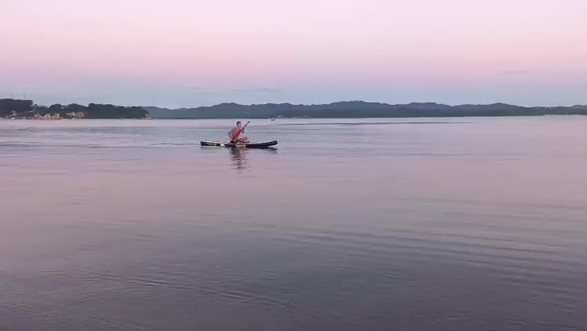 A man is paddleboarding on a calm body of water as the sun sets. The sky is a gradient of pink and purple. The man is wearing a pink shirt and is sitting on the paddleboard. He is holding a paddle in his hands and is in the process of paddling. The paddleboard is black. The water is calm and reflects the colors of the sky. In the distance, there is a shoreline with trees and hills. There are a few small boats in the water. The overall mood of the video is peaceful and serene.