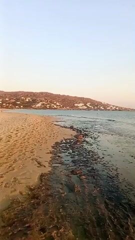 The video shows a scenic view of a beach with a shoreline. The sand is golden, and the water is a clear blue. The waves are gently lapping the shore. In the distance, there are hills with white buildings on them. The sky is a pale blue with a hint of pink, indicating either sunrise or sunset. There are no people or objects visible in the immediate vicinity of the beach. The scene is calm and tranquil.