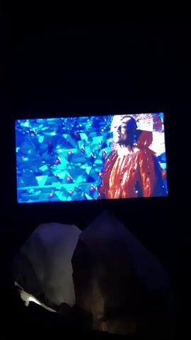 A screen displays a person wearing a decorative headdress and an orange garment. The background is a blurry mix of blue and green lights. Below the screen are two large, translucent, irregularly shaped objects that appear to be crystals. The scene is dark.