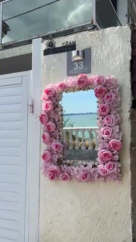 A close-up shot shows a decorative mirror on an exterior wall. The mirror is framed with artificial pink roses and seashells. Behind the mirror, a balcony railing and the sea are visible. A house number 33 with the street name "Primorska" is mounted above the mirror. To the left of the mirror is a white gate or door. The wall itself is textured and painted in a light color. The overall impression is of a whimsical, outdoor decoration.